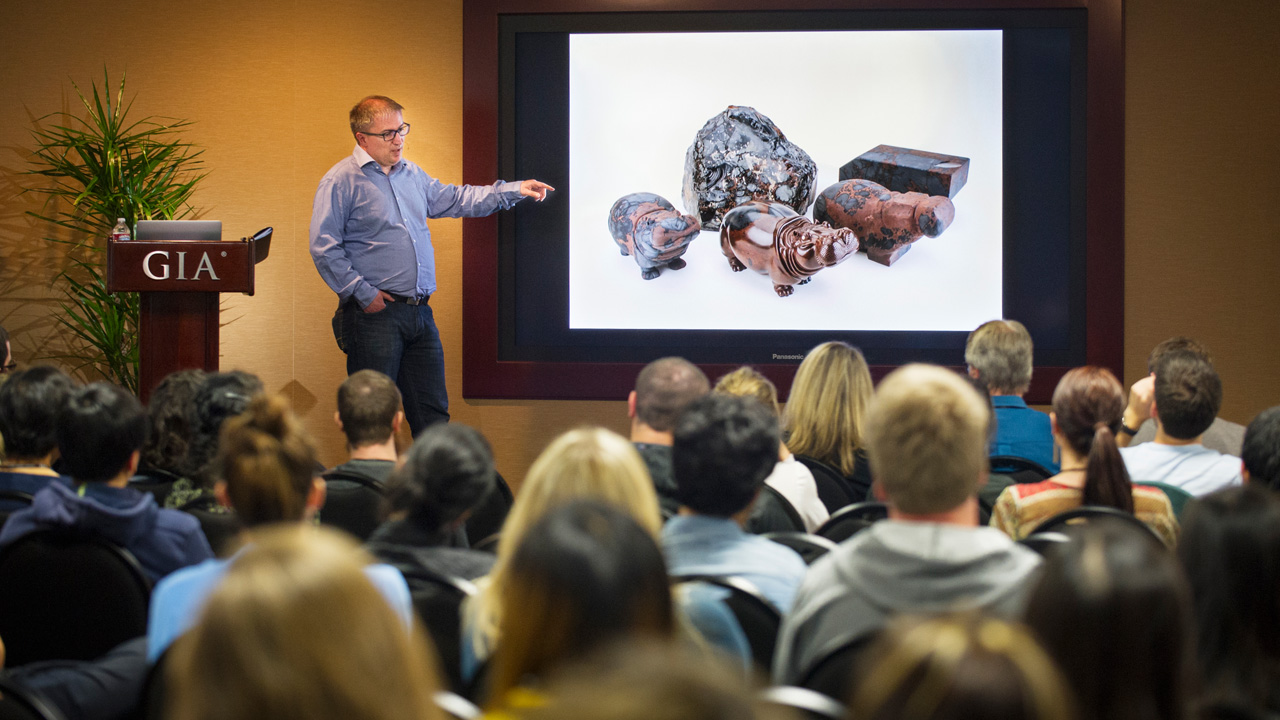 Dreher presented photos of his carvings alongside rough gems to show how he works through each piece to reveal the animal figure. The obsidian hippos pictured on the screen are  on display in Carlsbad as part of the “Generations of Mastery” exhibit. Photo by Kevin Schumacher/GIA