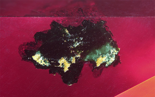 Figure 3. A cluster of dark green transparent malachite crystals breaks the crown of the cuprite. Photomicrograph by Britni LeCroy; field of view 2.81 mm.