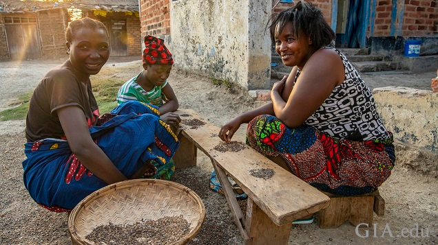 Three woman sit on the ground around a low bench/table to sort through gravel for gems.