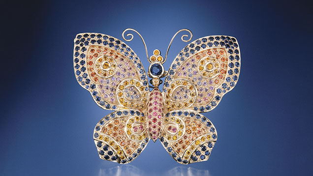 A colorful butterfly brooch adorned with various gemstones, featuring intricate designs and a shimmering finish against a blue background.