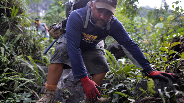 Diaz needs to crouch low as he makes his way through jungle terrain with a backpack on. He wears boots and gloves to protect him on the uncut path.